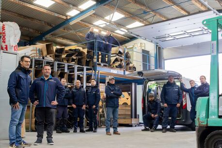 Das Team von Delewski steht nebeneinander in der Arbeitshalle. Im Hintergrund ein Firmenwagen.
