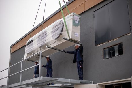 Monteure der Delewski Kälte- und Klimatechnik GmbH heben mit einem Kran ein großes verpacktes Kühlaggregat auf eine Metallplattform an der Außenwand eines modernen Gebäudes.