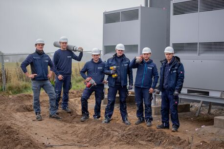 Sechs Monteure der Firma Delewski Kälte- und Klimatechnik GmbH mit Schutzhelmen posieren auf einer Baustelle vor einer modernen Industrie-Kühlanlage, ausgestattet mit Werkzeugen und Gasflaschen.