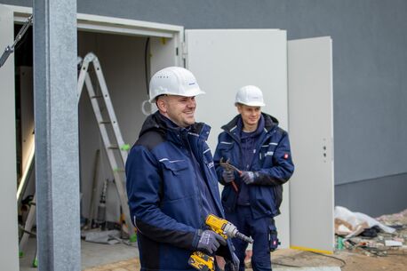 Zwei Monteure mit Schutzhelmen und Werkzeugen stehen bei Bauarbeiten vor einem Gebäude.