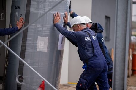 Zwei Monteure bringen gemeinsam mit einem Kollegen einen Wärmepumpe in ein Gebäude. Alle tragen Helme und Arbeitskleidung auf einer Baustelle.
