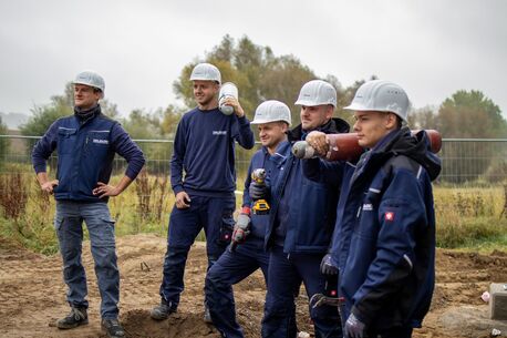 Fünf Bauarbeiter mit Schutzhelmen posieren lächelnd auf einer Baustelle im Freien, ausgestattet mit Werkzeugen und Arbeitskleidung.