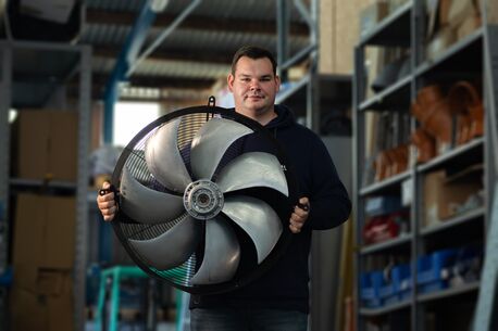 Ein Mitarbeiter hält einen großen Lüfter in seinen Händen und steht in der Werkstatthalle.