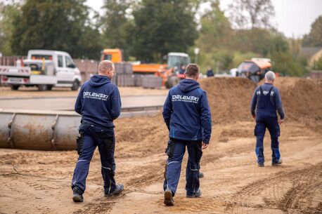 Drei Monteure der Firma Delewski Kälte- und Klimatechnik GmbH gehen auf einer Baustelle über den Sandboden. Im Hintergrund befinden sich Baufahrzeuge und Baumaterial.