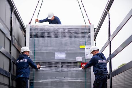 Drei Monteure positionieren mit Schutzhelmen eine große industrielle Kühlanlage mithilfe eines Krans auf einem LKW.